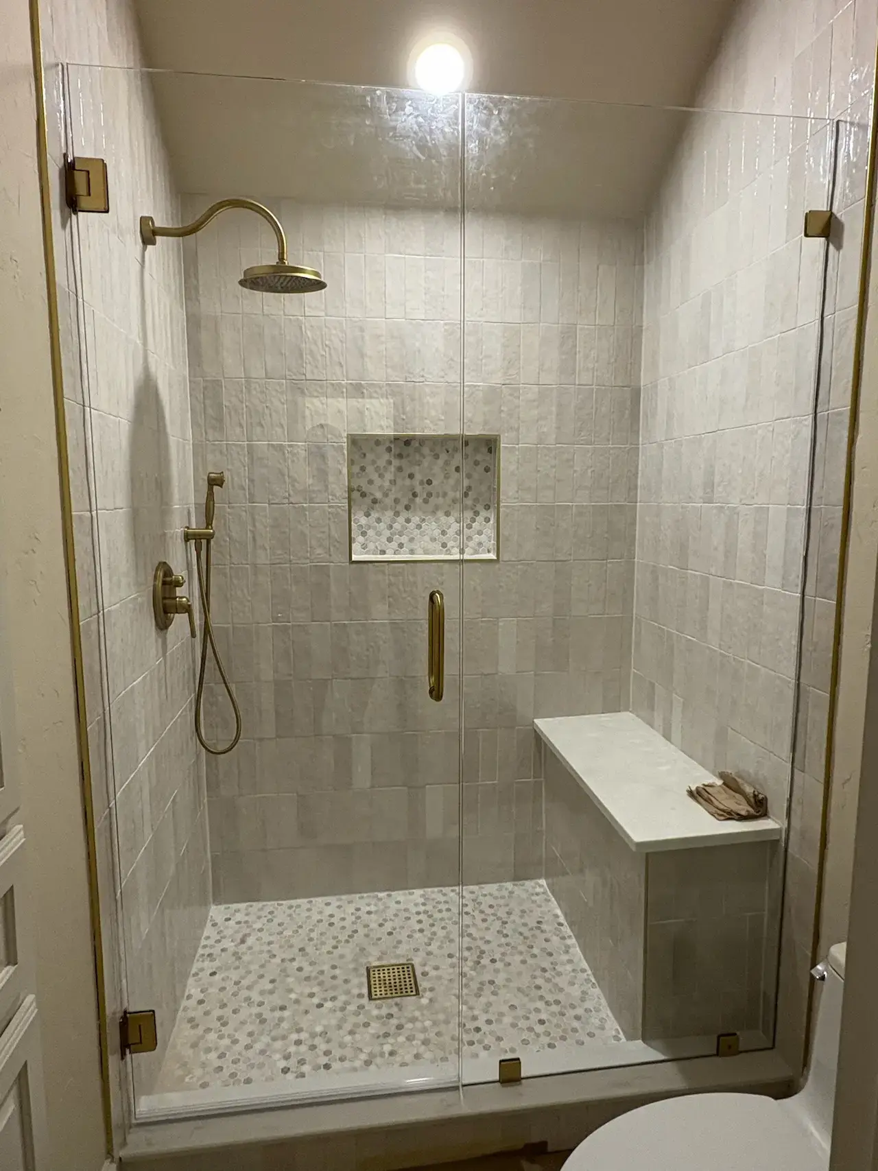 A brightly and warmly lit shower with a borderless glass door, with small cream-colored tiles and matte bronze colored shower head, handles, hinges, and handles with a bench inside